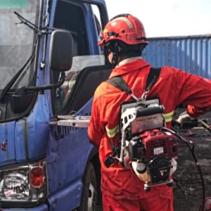 A rescue operator wearing a heavy-duty backpack gasoline hydraulic pump while operating a connected hydraulic combi tool to cut and spread a crushed car frame at an accident scene. (ภาพเจ้าหน้าที่กู้ภัยสวมชุดปั๊มไฮดรอลิกเครื่องยนต์เบนซินแบบสะพายหลัง พร้อมกำลังใช้เครื่องมือตัด-ถ่างอเนกประสงค์ที่ต่อพ่วงกัน ทำการตัดและถ่างโครงสร้างตัวถังรถยนต์ที่พังยับเยินในจุดเกิดเหตุ)