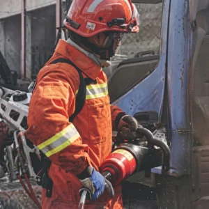 A close-up shot of a hydraulic combi tool shearing through a reinforced car B-pillar, connected via a high-pressure hose to a gasoline pump worn on a rescuer's backpack. (ภาพมุมใกล้ของเครื่องมือ Combi Tool กำลังตัดเสาบีเหล็กกล้าของรถยนต์ โดยมีสายไฮดรอลิกแรงดันสูงเชื่อมต่อไปยังปั๊มเบนซินที่สะพายอยู่บนหลังของเจ้าหน้าที่กู้ภัย)