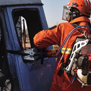 Side view of a firefighter wearing an ergonomic padded backpack gasoline hydraulic power pack while forcing open a jammed vehicle door with a combi tool. (ภาพด้านข้างของเจ้าหน้าที่ดับเพลิงสวมชุดปั๊มไฮดรอลิกเบนซินสะพายหลังบุนุ่มหนาพิเศษ กำลังใช้เครื่องมือ Combi Tool งัดเปิดประตูรถยนต์ที่ติดขัด)